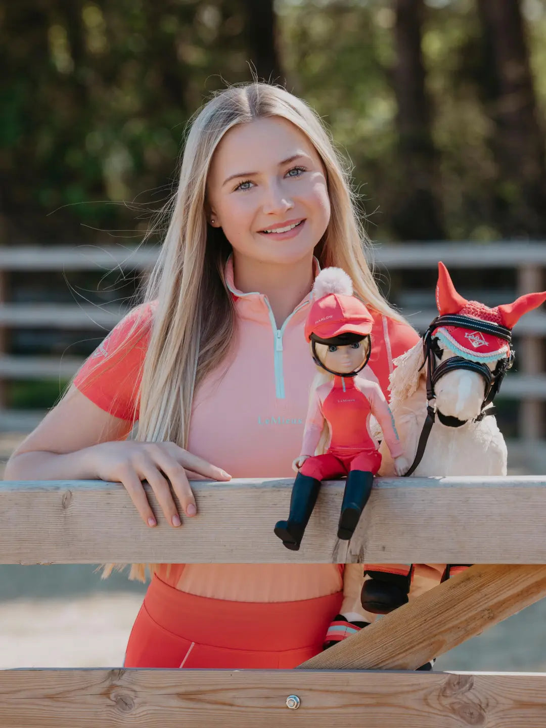 Front view of a young model standing behind a harlow doll who is sitting on the fence, while the model is holding a palomino toy pony with a coral harlow fly hood