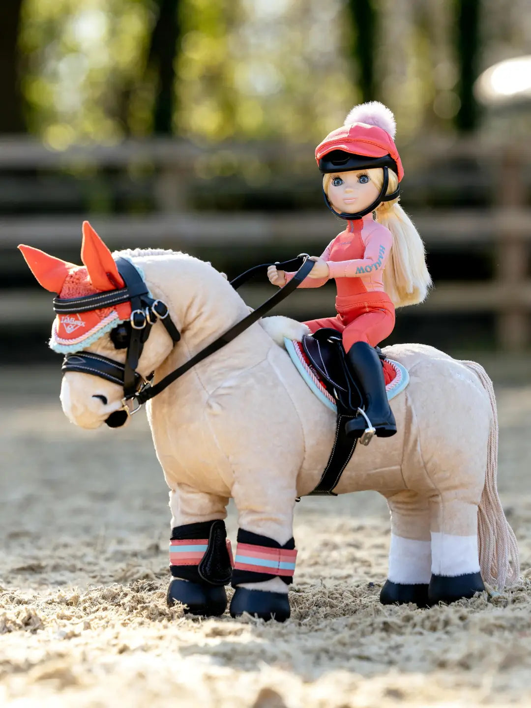 Side view of a doll riding a toy pony that's wearing the coral LeMieux Harlow Toy Pony Pad and matching accessories while standing in an outdoor arena