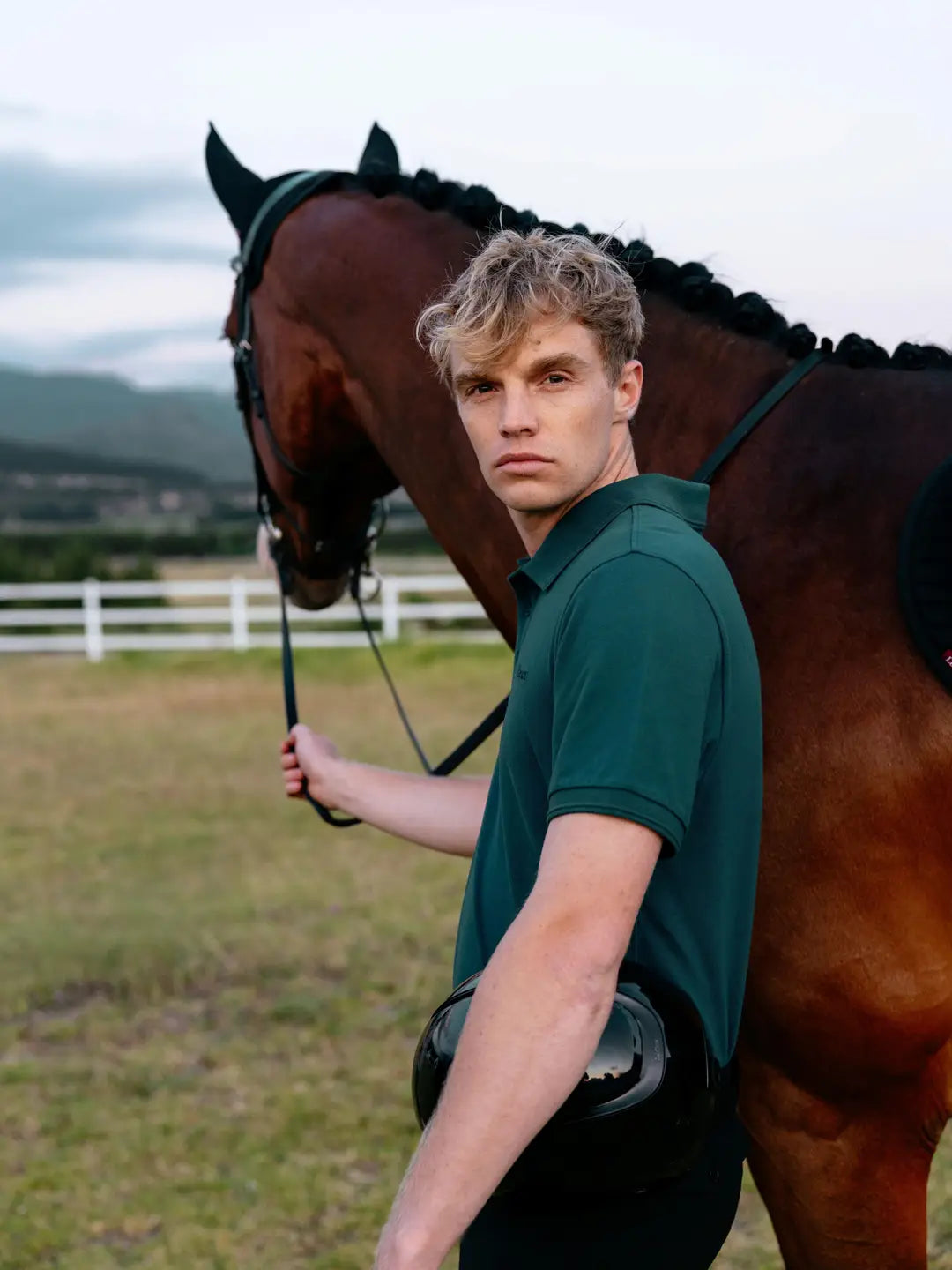 Man wearing LeMieux Men’s Classique Polo Shirt in Jungle green standing beside a horse and the horse's left rein outdoors