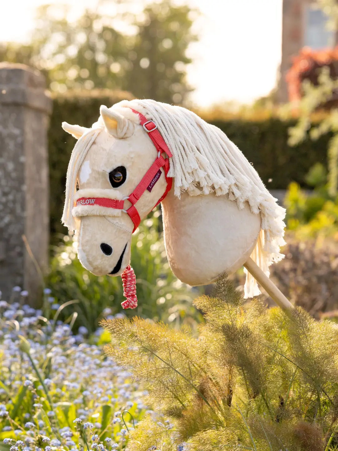 Side view of a palomino hobby horse wearing a coral LeMieux Hobby Horse Harlow Headcollar & Leadrope with a garden background