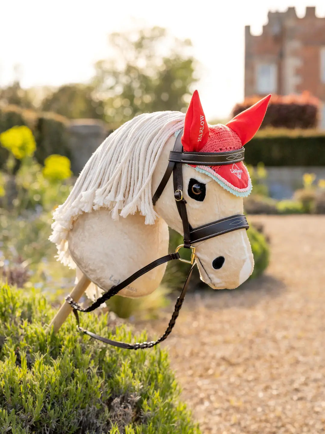 Angled front view of a palomino hobby horse wearing the coral LeMieux Hobby Horse Harlow Fly Hood in a garden setting