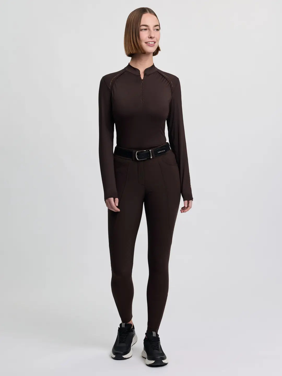 Full body front view of a model wearing the brown LeMieux Brilliance Knee Grip Breeches, brown long sleeve top, and black tennis shoes on a plain background