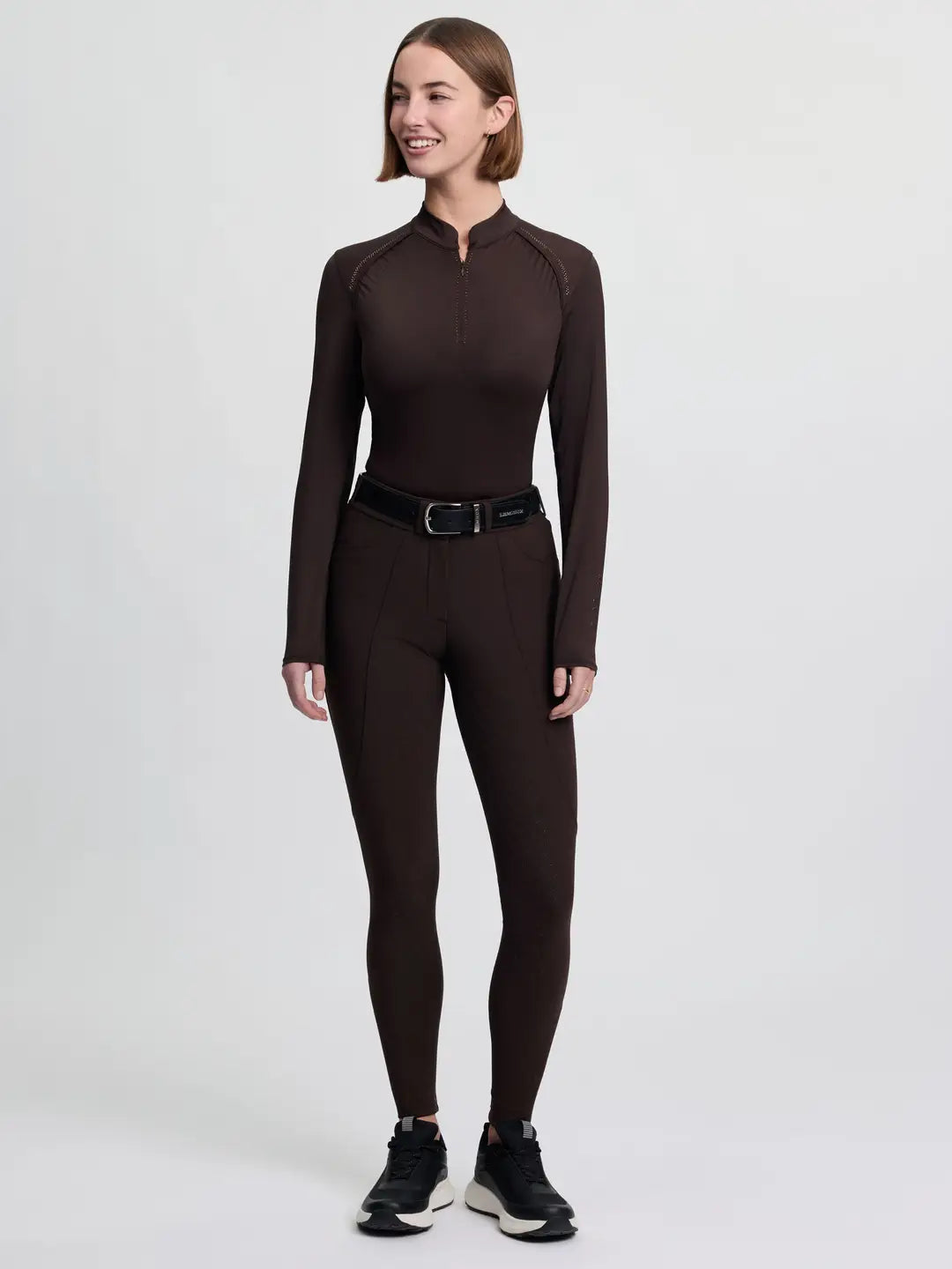 Full body front view of a model wearing the brown LeMieux Brilliance Full Seat Breeches, brown long sleeve shirt, and black tennis shoes on a plain background