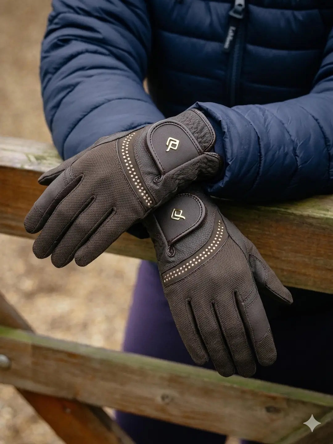 Front view of a model standing against a fence with arms crossed at the wrists while wearing a pair of brown LeMieux Crystal Gloves 