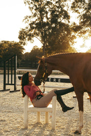 Side view of a female model wearing the cherry blossom Criniere Kai Shirt while sitting in a chair in an arena with a horse in front of her 
