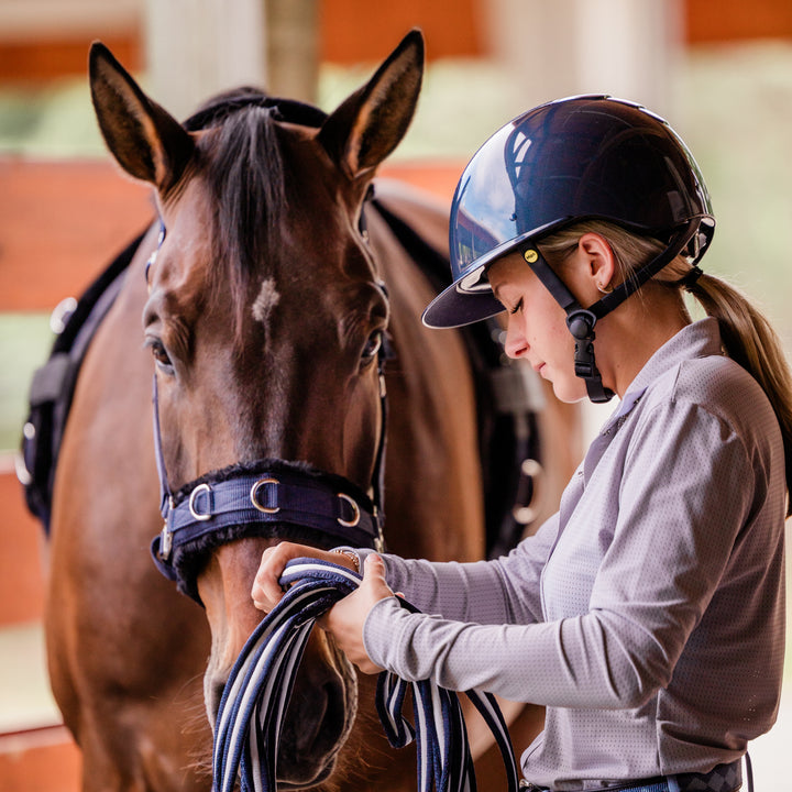 Side view of a female model wearing the Navy/Navy Shimmer Equinavia Crown Glossy MIPS Riding Helmet while standing next to a bay horse
