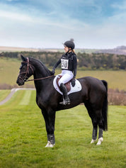 Side view of a woman wearing the black LeMieux Eventing Bib while sitting on a black horse in a green field