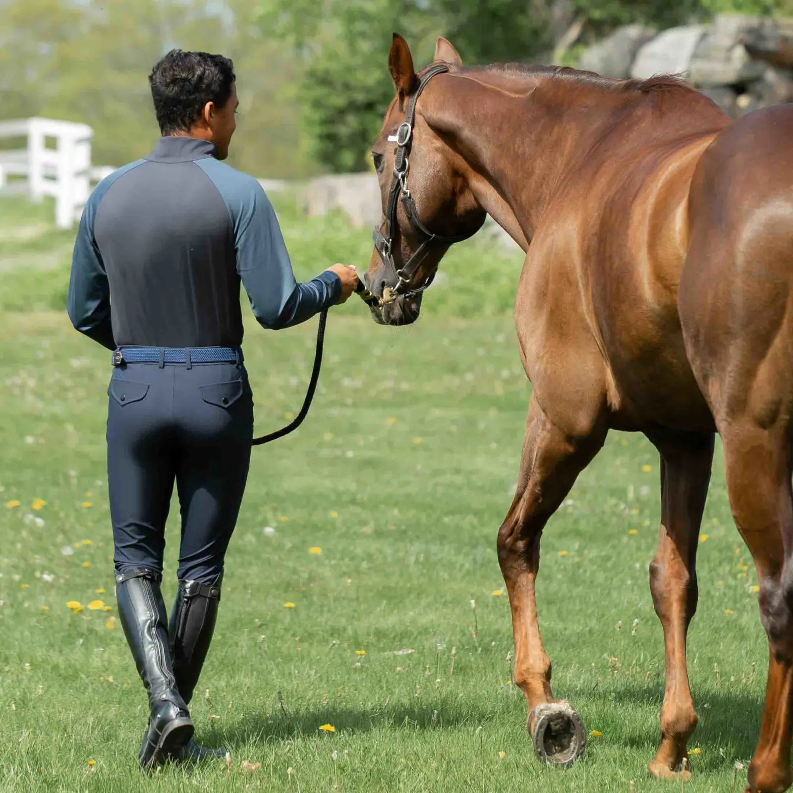 Back view of a man leading a horse while wearing the navy Equinavia Karl Men's Knee Patch Breeches, matching shirt, and black tall boots