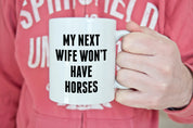 Front view of a model holding the Canary Road My Next Wife Coffee Mug  while the model is wearing a pink sweatshirt