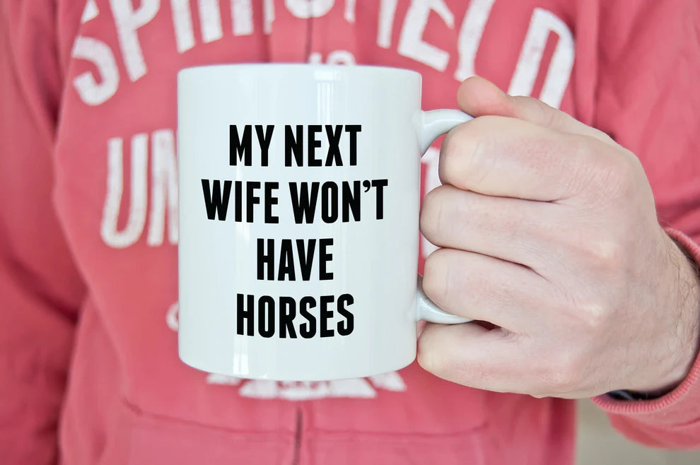 Front view of a model holding the Canary Road My Next Wife Coffee Mug  while the model is wearing a pink sweatshirt