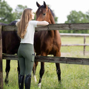 Back view of a female model wearing the Beetle Khaki Green Horze Gillian Silicone Full Seat Tights while leaning on a fence with a horse standing directly behind it