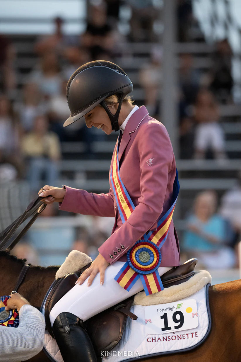 Side view of a woman in a pink show coat sitting on a horse with a white saddle pad and 2 Pink Bow Dubois Cheval Number Pin Charms on the number 19
