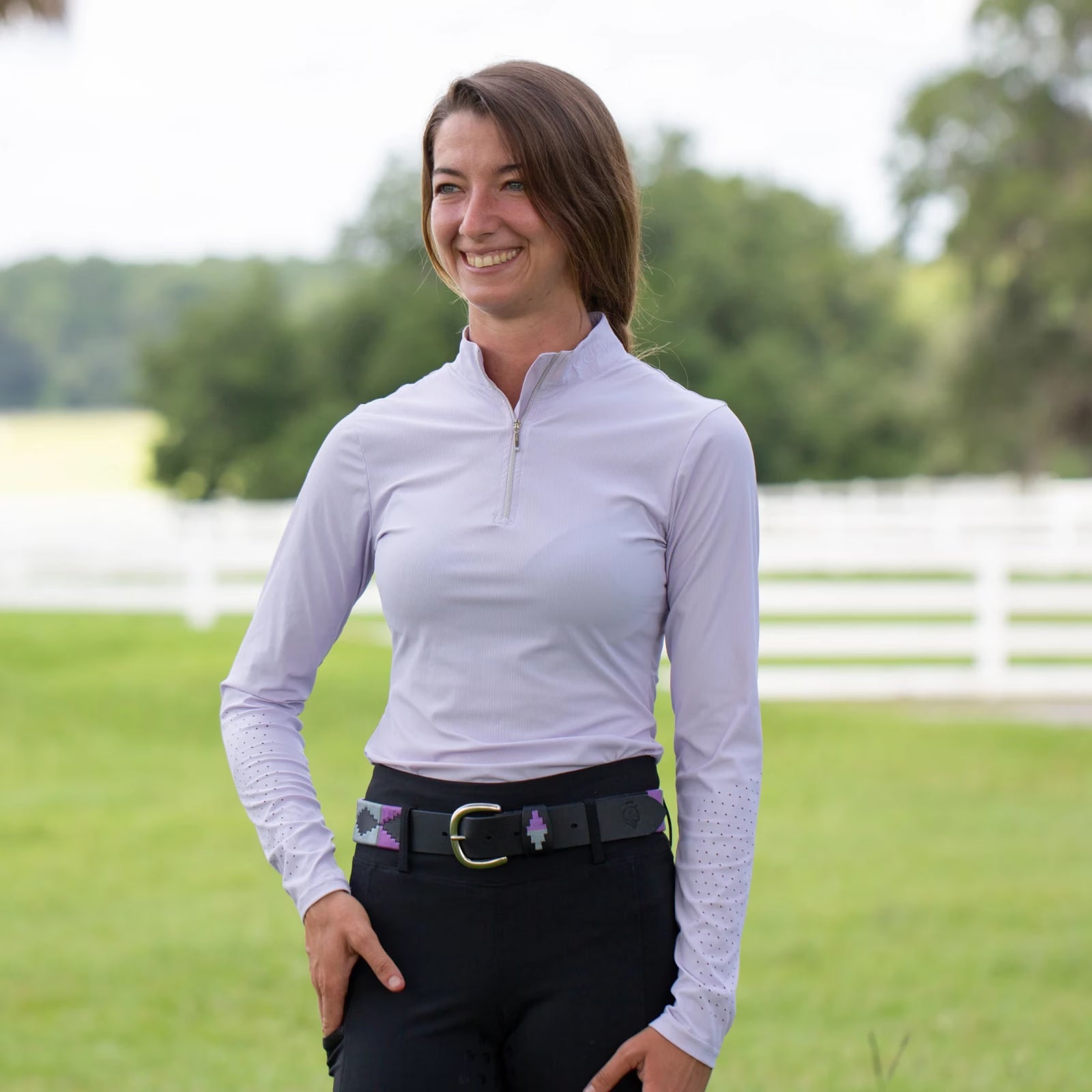 Front view of a model standing outside wearing a purple top and the purple and gray, black-leather Equinavia Siri Polo Belt on black breeches