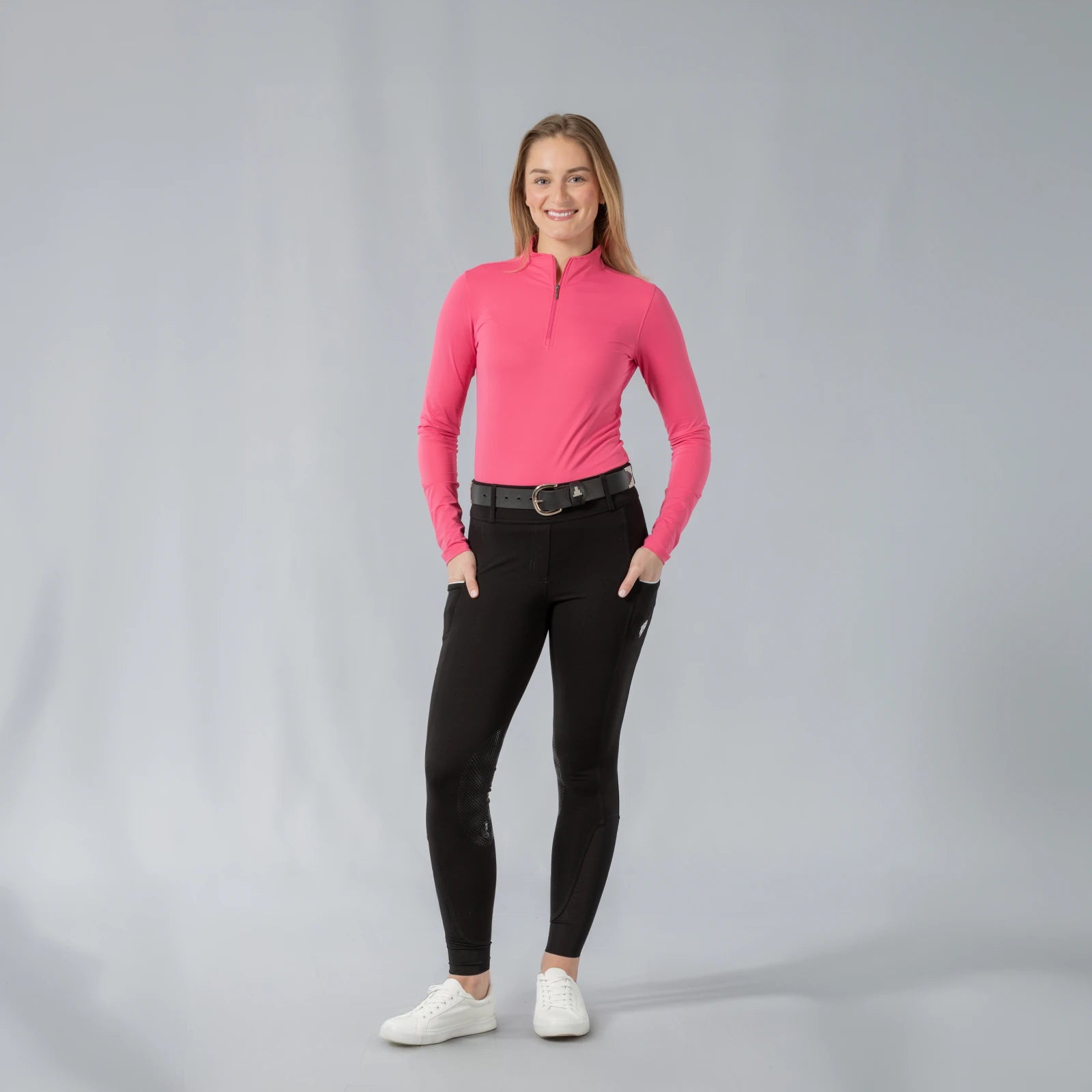 Full body front view of a model wearing the Equinavia Kari Womens Long Sleeve Sun Shirt with NordicAir Tech in pink, black breeches, and white sneakers on a gray background