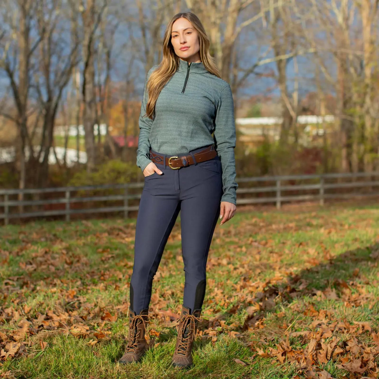 Front view of a woman standing outside wearing a green top, blue breeches, and the navy brown-leather Equinavia Siri Polo Belt
