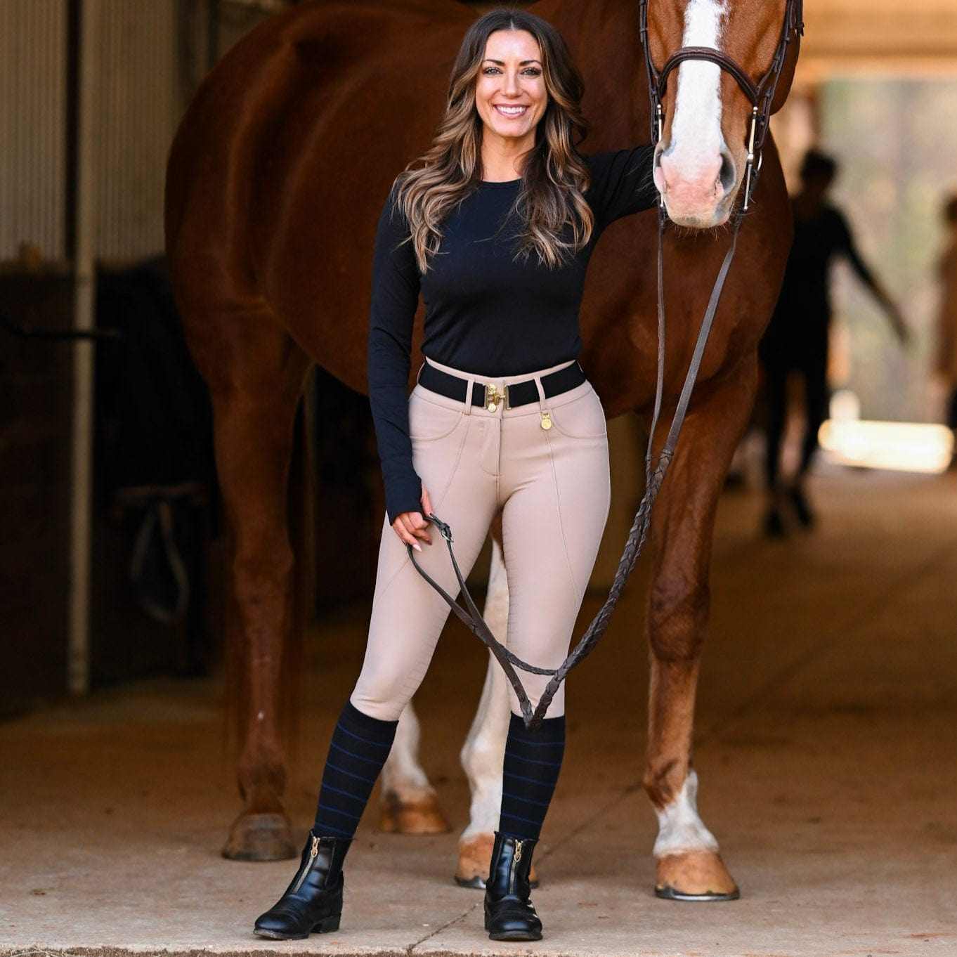 Full body front view of a female model wearing the black with navy stripe Dreamers & Schemers Simple Stripe Knit Boot Socks while standing in front of a chestnut horse in a barn aisle