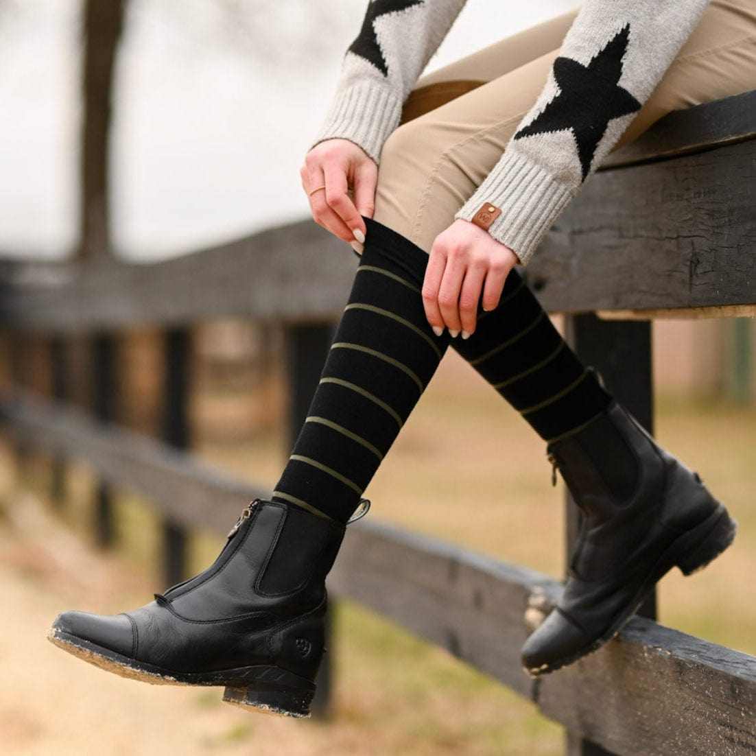 Close-up side view of a model wearing the black with sage stripe Dreamers & Schemers Simple Stripe Knit Boot Socks while sitting on a black wooden fence