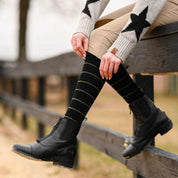 Close-up side view of a model wearing the black with sage stripe Dreamers & Schemers Simple Stripe Knit Boot Socks while sitting on a black wooden fence