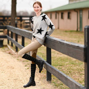 Full body view of a model wearing the black with sage stripe Dreamers & Schemers Simple Stripe Knit Boot Socks with tan breeches and gray sweater while leaning up against a black wooden fence