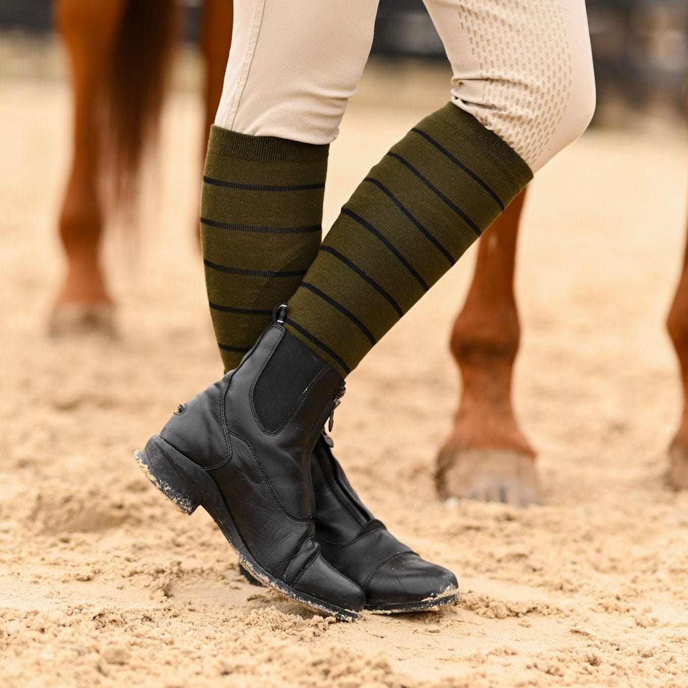 Side view of a model wearing the sage with black stripe Dreamers & Schemers Simple Stripe Knit Boot Socks while standing in an arena