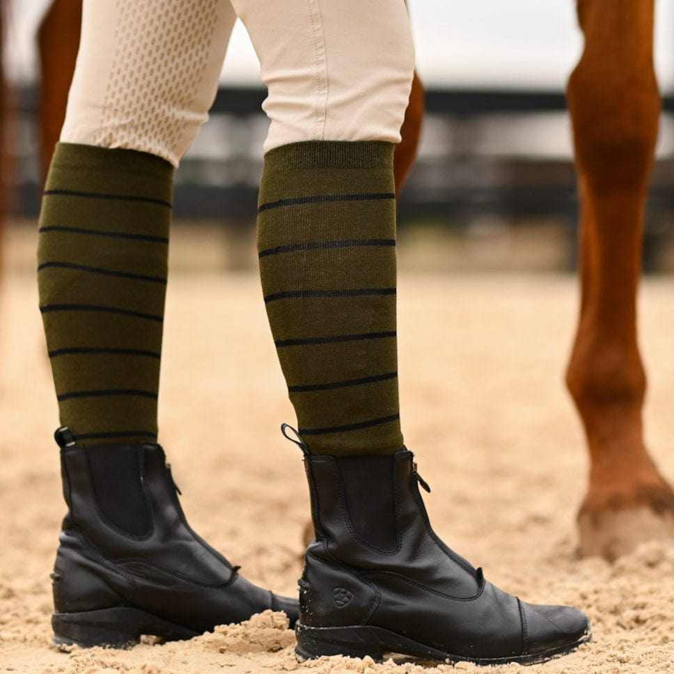 Side view of a model wearing the sage with black stripe Dreamers & Schemers Simple Stripe Knit Boot Socks while standing in an arena