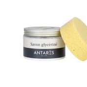 Front view of a jar of Antares Savon Glycerine with a yellow sponge leaned up against it on a white background