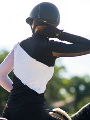 Back view of a female model wearing the black and white Criniere Sophie Stride & Conquer Schooling Shirt while riding a horse