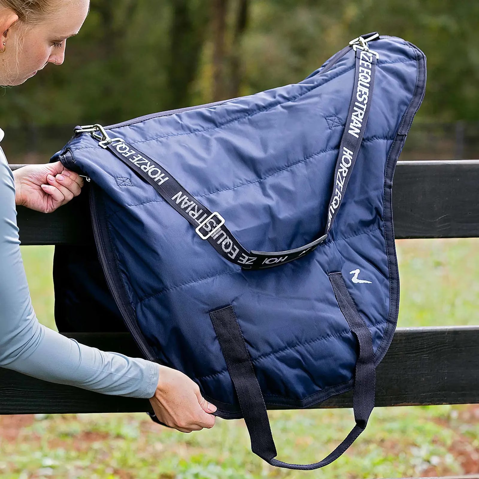 Side view of the navy Horze Saddle Bag sitting on a black fence outside while a woman is unzipping the bag
