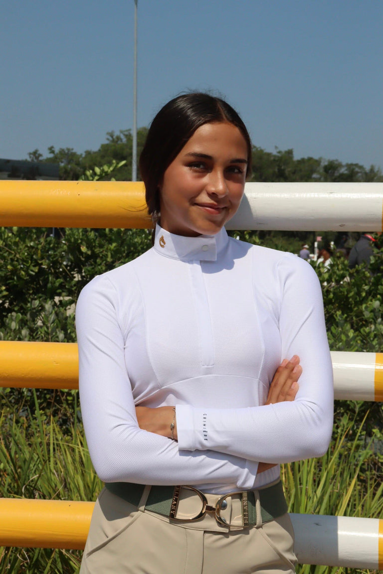 Front view of a female model wearing the white Criniere Monique The Winner's Edge Mesh Show Shirt while standing in front of a horse jump