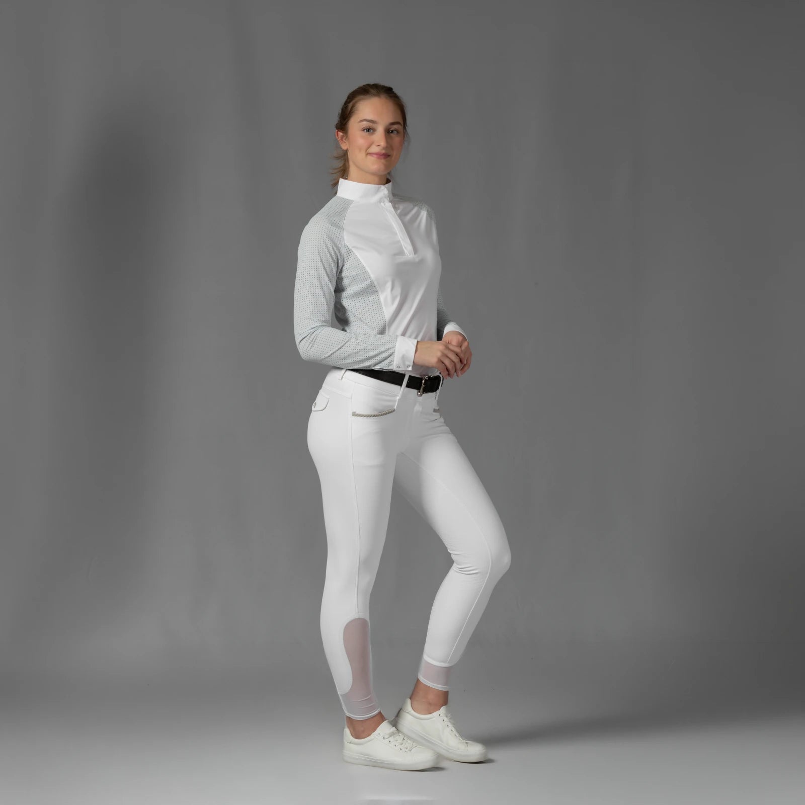 Full body front view of a model wearing the Equinavia Sara Womens Perforated Long Sleeved Show Shirt in light blue and white, white breeches, and white sneakers on a gray background