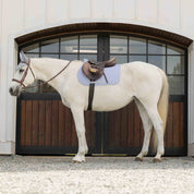 Full side view of a gray horse standing in front of a barn while wearing the gray Equinavia Harstad All Purpose Pad under a brown saddle