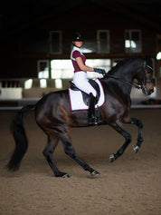Side view of a female model riding a dark colored horse with a bordeaux  Equestrian Stockholm White Perfection Dressage Pad