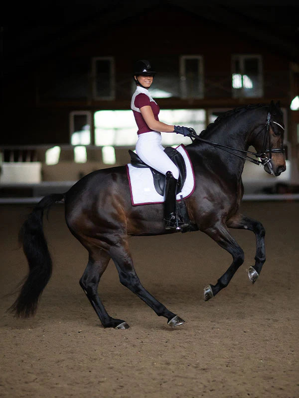 Side view of a female model riding a dark colored horse with a bordeaux  Equestrian Stockholm White Perfection Dressage Pad