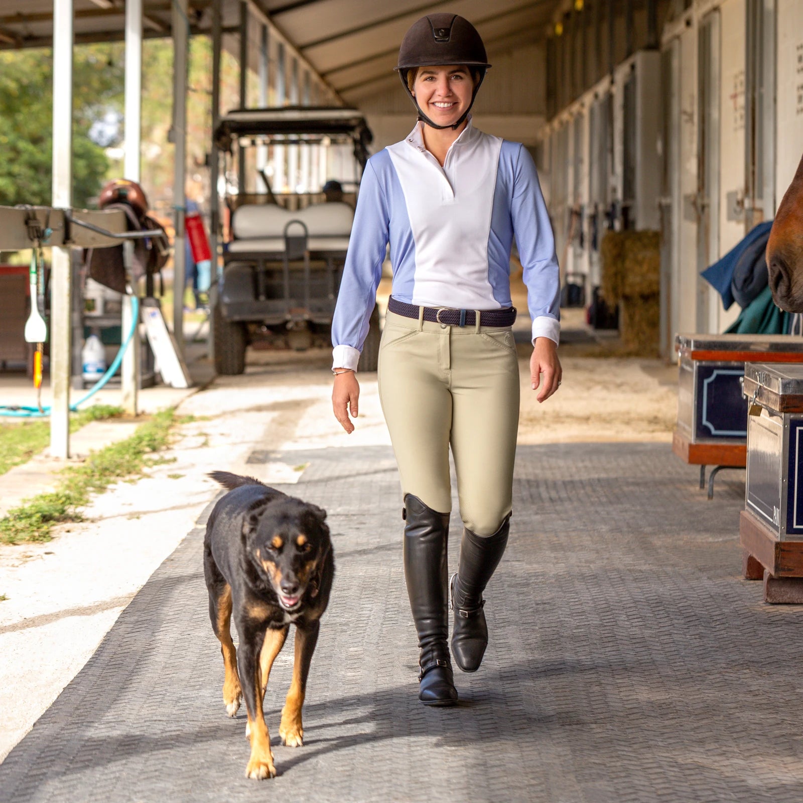 Front view of a model wearing the tan Equinavia Maud Knee Patch Breeches, blue and white top, and black tall boots while walking next to a dog down a barn aisle