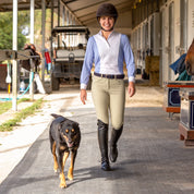 Front view of a model wearing the tan Equinavia Maud Knee Patch Breeches, blue and white top, and black tall boots while walking next to a dog down a barn aisle