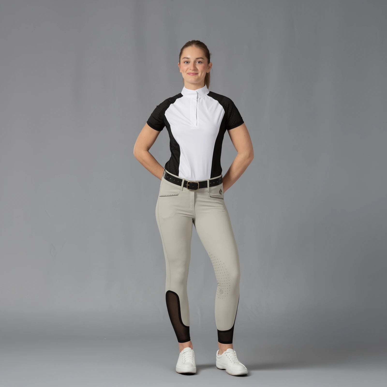 Full body front view of a model wearing the Equinavia Sara Womens Perforated Short Sleeved Show Shirt in black and white, tan breeches, and white sneakers on a gray background