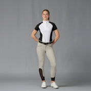 Full body front view of a model wearing the Equinavia Sara Womens Perforated Short Sleeved Show Shirt in black and white, tan breeches, and white sneakers on a gray background
