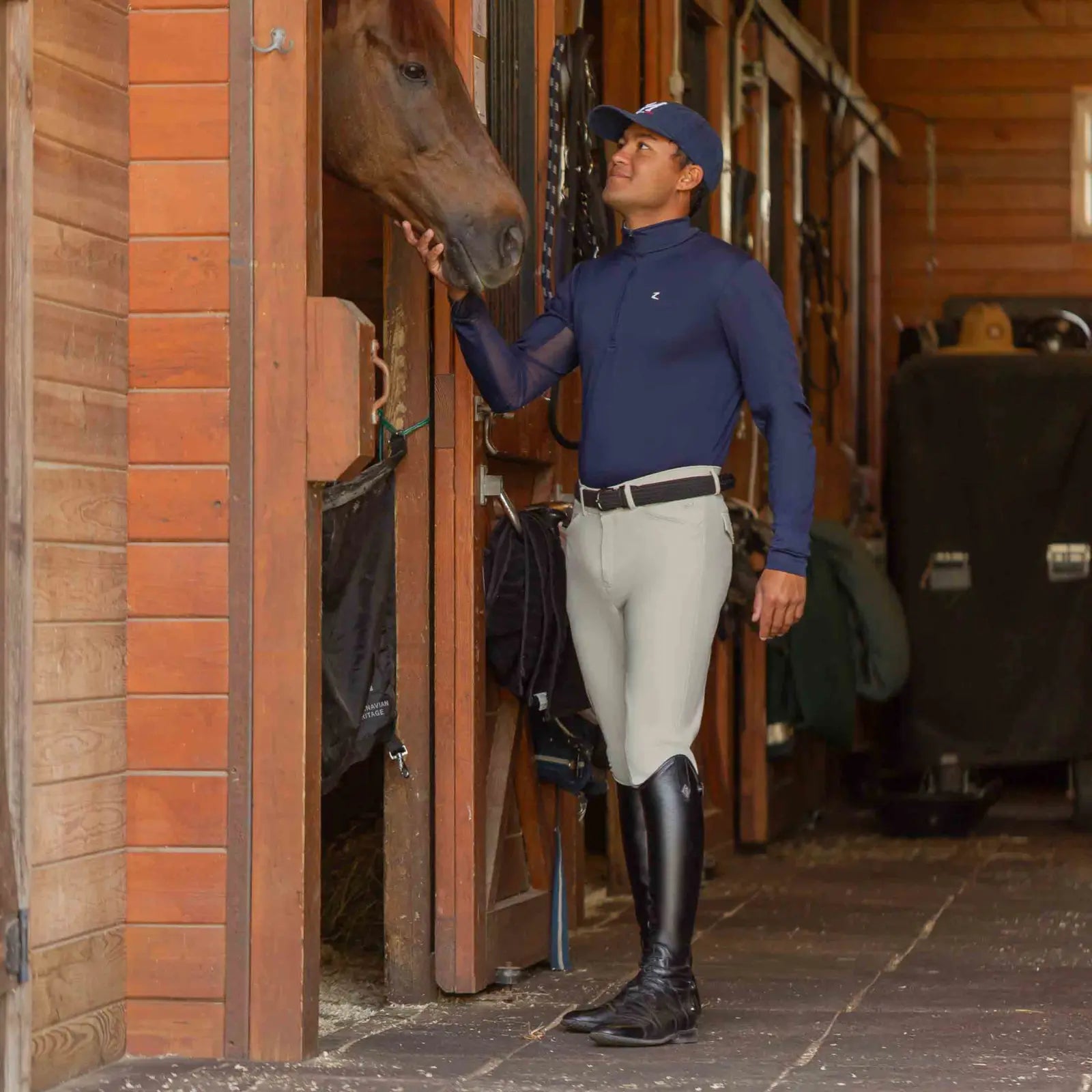 Angled front view of a model wearing the tan Equinavia Anders Mens Full Seat Breeches while standing in a barn petting a horse in a stall